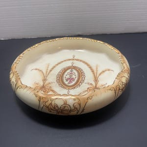 Puede incluir: Un cuenco redondo de cerámica color crema con borde festoneado y ribete dorado. El cuenco presenta un diseño ovalado central con motivos florales y de lazo en tonos rosa, marrón y dorado. Adornos de volutas decorativas adornan el exterior del cuenco.