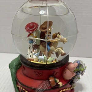 May include: A Toy Story-themed snow globe featuring Woody, Buzz Lightyear, and other characters inside a clear glass globe. The base is red with gold accents and a green, textured side.