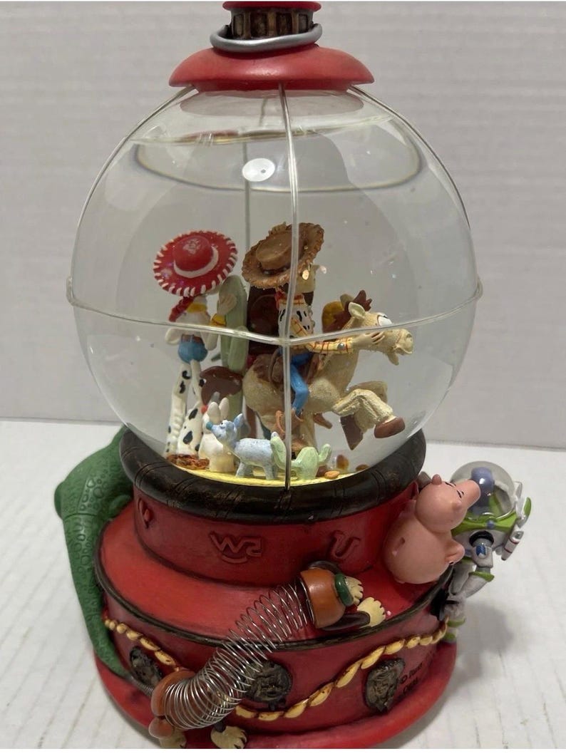 May include: A Toy Story-themed snow globe featuring Woody, Buzz Lightyear, and other characters inside a clear glass globe. The base is red with gold accents and a green, textured side.