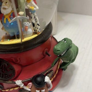 May include: A Toy Story-themed snow globe featuring multiple characters. The base is red with gold accents, and a green dinosaur figure is attached to the side. Inside the globe are figures of Woody, Jessie, and other characters.