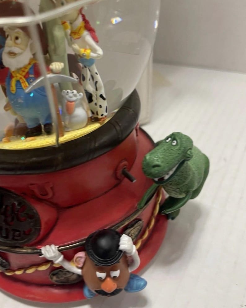 May include: A Toy Story-themed snow globe featuring multiple characters. The base is red with gold accents, and a green dinosaur figure is attached to the side. Inside the globe are figures of Woody, Jessie, and other characters.