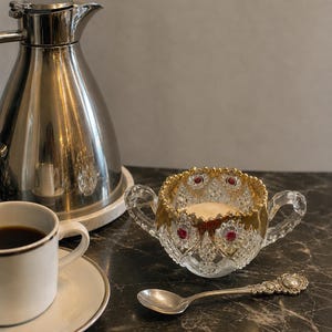 May include: A clear cut glass sugar bowl with two handles, featuring gold-painted scalloped rims and embedded red circular accents. The bowl is accompanied by a small silver-toned decorative spoon with an ornate handle design.