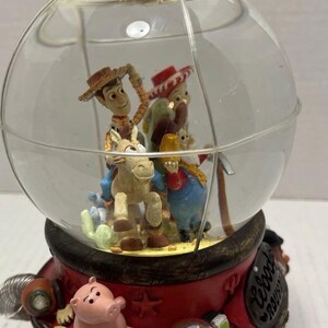 May include: A Toy Story-themed snow globe featuring Woody riding a horse, with other characters inside the glass globe. The base is red with additional characters, including a pig and Buzz Lightyear. The globe is topped with a red cap.