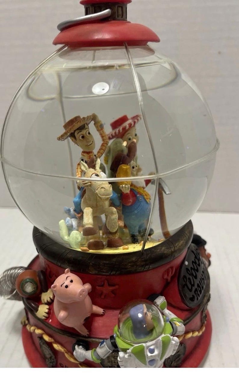 May include: A Toy Story-themed snow globe featuring Woody riding a horse, with other characters inside the glass globe. The base is red with additional characters, including a pig and Buzz Lightyear. The globe is topped with a red cap.