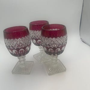 Może przedstawiać: Trzy kieliszki do wina w stylu vintage z rubinowoczerwonymi obwódkami i miseczkami z diamentowym wzorem. Kieliszki mają przezroczyste, kwadratowe podstawy i nóżki. Kieliszki są prawdopodobnie wykonane z kryształu lub podobnego materiału.