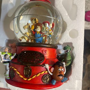 May include: A Toy Story themed snow globe featuring characters inside a clear glass globe. The base is red with gold accents and the words "Woody's Roundup". Characters include Woody, Buzz Lightyear, and Mr. Potato Head.