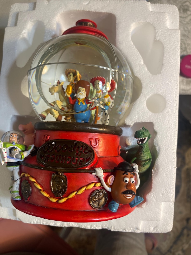 May include: A Toy Story themed snow globe featuring characters inside a clear glass globe. The base is red with gold accents and the words "Woody's Roundup". Characters include Woody, Buzz Lightyear, and Mr. Potato Head.