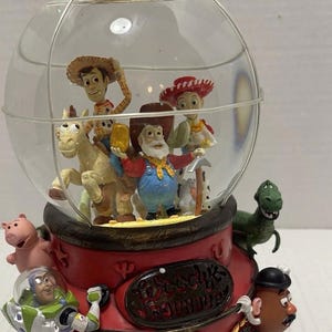 May include: A red and clear Toy Story-themed snow globe. Inside the globe are figurines of Woody, Jessie, and other characters. The base is red with the words "Woody's Roundup" and other character figurines.