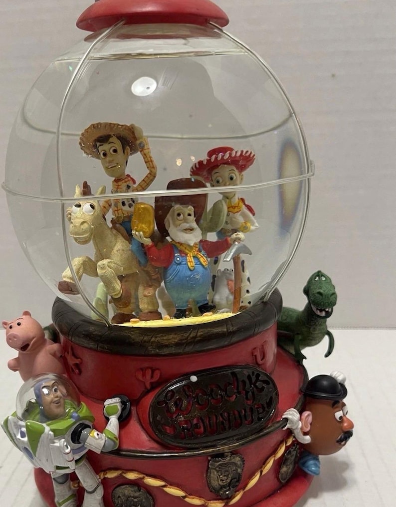 May include: A red and clear Toy Story-themed snow globe. Inside the globe are figurines of Woody, Jessie, and other characters. The base is red with the words "Woody's Roundup" and other character figurines.