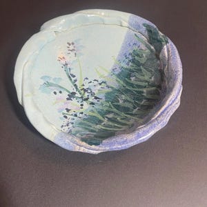 May include: A handmade ceramic bowl with a light blue interior and a textured, uneven rim. The bowl features a painted floral and landscape design in shades of green, purple, and black. The bowl is approximately 6 inches in diameter.