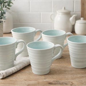 Puede incluir: Cinco tazas de cerámica blanca con interior azul claro y diseño exterior acanalado. Las tazas están dispuestas sobre una superficie de madera con un paño de color neutro. Una tetera, un azucarero y una tabla de cortar están en el fondo.