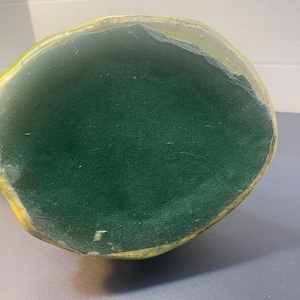 Pode incluir: Um objeto oval verde com um interior verde escuro e aveludado. O exterior tem uma borda amarela e dourada. O objeto est&aacute; sobre uma superf&iacute;cie cinza.