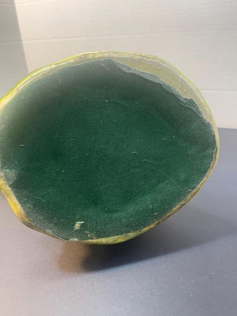 Pode incluir: Um objeto oval verde com um interior verde escuro e aveludado. O exterior tem uma borda amarela e dourada. O objeto est&aacute; sobre uma superf&iacute;cie cinza.