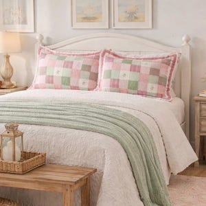 May include: A bedroom scene featuring a white bed with pink and green checkered pillows and a light green throw blanket. Two wooden bedside tables with lamps flank the bed. A wooden bench with a woven tray and lantern sits at the foot of the bed.