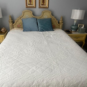 May include: A white bedspread with intricate quilted patterns covering a bed. The bed features a light yellow headboard with floral accents, two blue square pillows, and is flanked by two matching yellow nightstands with lamps and decor.