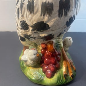 Pode incluir: Estatueta de galinha de cer&acirc;mica com corpo salpicado de branco e preto, pousada sobre uma base verde com um cacho de uvas vermelhas e um pintinho. As patas da galinha s&atilde;o laranja e pretas.
