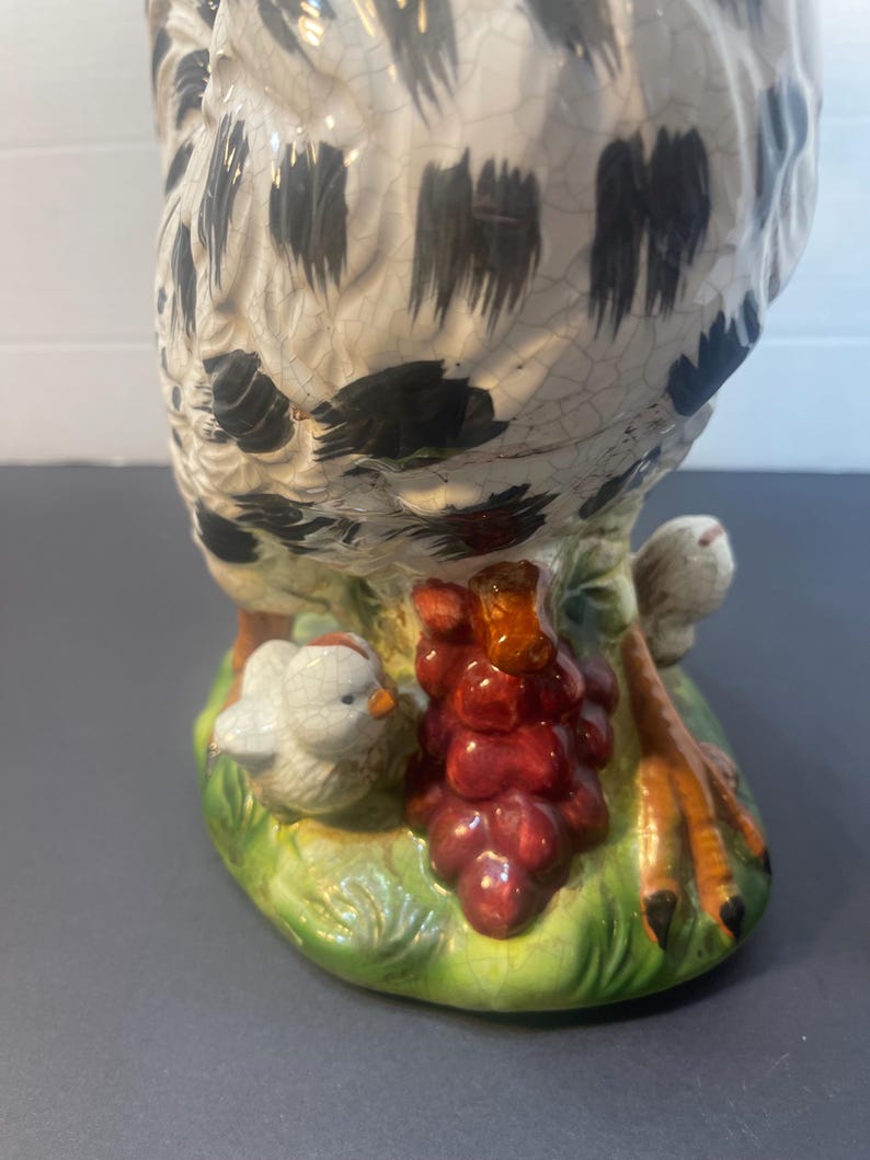 Pode incluir: Estatueta de galinha de cer&acirc;mica com corpo salpicado de branco e preto, pousada sobre uma base verde com um cacho de uvas vermelhas e um pintinho. As patas da galinha s&atilde;o laranja e pretas.