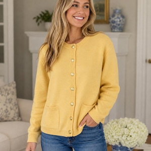 May include: A long-sleeved yellow knit cardigan featuring a round neckline, a front button closure with six metallic gold buttons, and two front patch pockets. The garment is paired with blue denim jeans, creating a casual and functional outfit.
