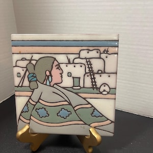 May include: Square ceramic tile with a Native American design. The tile features a woman in profile, wearing a gray shawl and turquoise earrings, set against a backdrop of pueblo-style buildings. The tile is displayed on a gold-colored stand.
