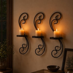 May include: Three black metal wall-mounted candle sconces with scrolling designs, each featuring a circular platform holding a single lit pillar candle with a warm glow, mounted vertically in a row on a plain wall surface.