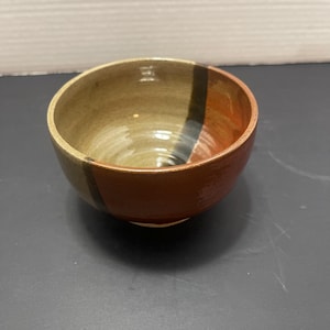 Puede incluir: Un cuenco de cerámica hecho a mano con forma redonda. El cuenco presenta un diseño de bloques de color con tonos de marrón, beige y naranja. Una franja vertical de negro y azul está en el lateral. El interior tiene un patrón en espiral.