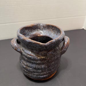 May include: A small, handmade ceramic pot with a textured, dark blue and brown exterior. The pot has two small handles and a slightly irregular rim. The interior is a lighter blue. The pot is likely used for decorative purposes.