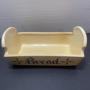 May include: Cream-colored ceramic bread basket with curved handles. The word "Bread" is written in black script across the front. The basket has a rectangular shape and is designed for serving bread.
