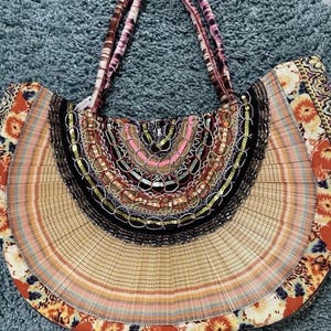 Peut inclure: Un grand sac fourre-tout coloré avec un design semi-circulaire. Le sac présente une variété de motifs et de textures, notamment des imprimés floraux, des sections tissées et des embellissements perlés. Les anses sont rayées de tons roses et bruns.