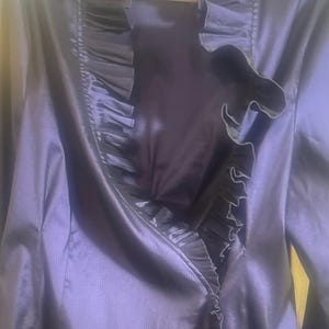 May include: A dark purple jacket with a ruffled trim. The jacket has long sleeves and a satin-like finish. The ruffled trim is a darker shade of purple and runs along the front opening and collar.