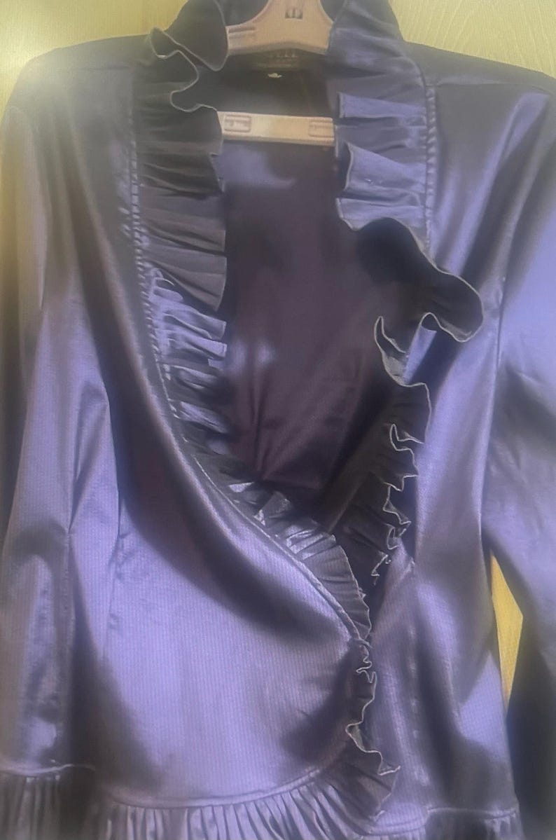 May include: A dark purple jacket with a ruffled trim. The jacket has long sleeves and a satin-like finish. The ruffled trim is a darker shade of purple and runs along the front opening and collar.