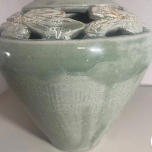 May include: A pale green ceramic urn with a lid. The urn has a bulbous body and a lid with floral cutouts. The floral cutouts are white. The urn is a decorative item.