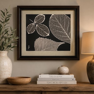 May include: A framed artwork featuring detailed leaf skeletons against a black background. The leaves are in shades of white and gray, with a dark brown frame. The artwork is displayed on a wooden surface.