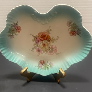 May include: An antique, scalloped-edge serving dish with a light blue rim and a floral design. The dish features a central bouquet of pink, orange, and yellow roses, with smaller floral arrangements around the edges. It is displayed on a gold-colored stand.