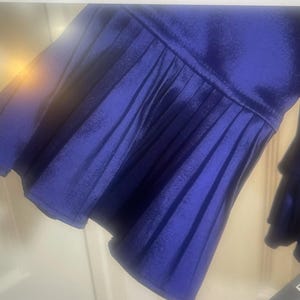 May include: Close-up of a royal blue garment sleeve with pleated detailing. The fabric has a slight sheen, and the sleeve flares out into a ruffled cuff. The garment appears to be made of a soft, flowing material.