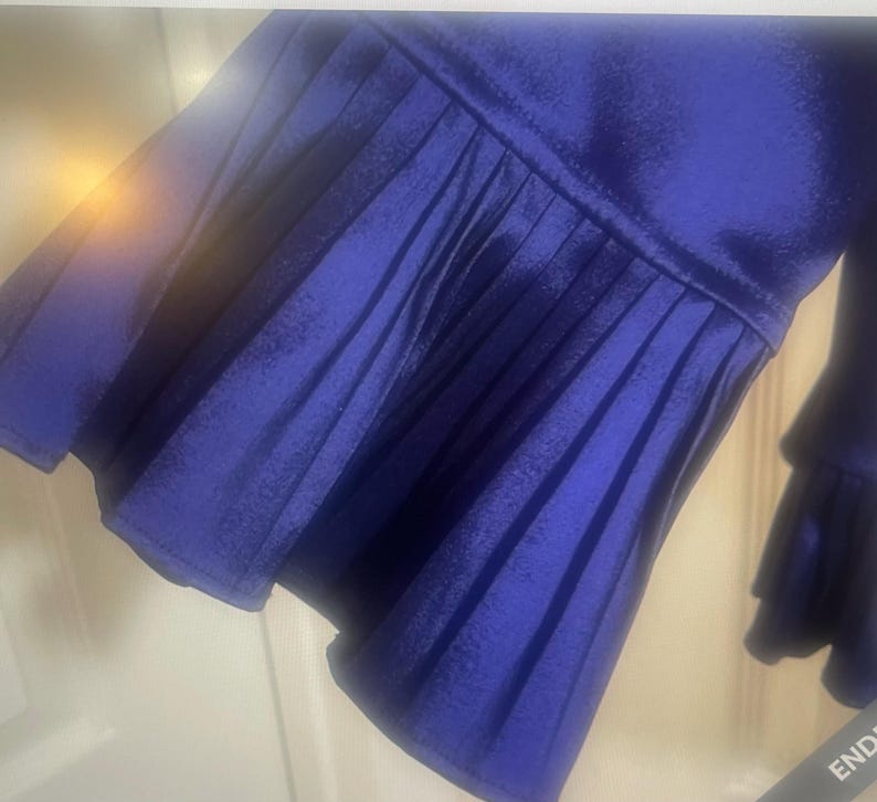 May include: Close-up of a royal blue garment sleeve with pleated detailing. The fabric has a slight sheen, and the sleeve flares out into a ruffled cuff. The garment appears to be made of a soft, flowing material.