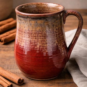 Peut inclure: Une tasse en céramique faite à la main, aux couleurs riches et terreuses. La tasse passe d'une base rouge profond à des tons bruns et beiges chauds sur le bord. Des bâtons de cannelle sont disposés sur une surface en bois.