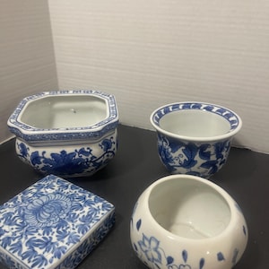 May include: Four white ceramic containers with blue floral patterns. The collection includes a hexagonal bowl, a round bowl, a small box, and a rounded container. The designs feature intricate floral and leaf motifs.