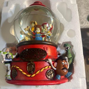 May include: A Toy Story themed snow globe featuring Woody, Buzz Lightyear, Mr. Potato Head, Rex, and other characters. The base is red with gold accents and the text "Woody's Roundup". The globe is clear with a red top.
