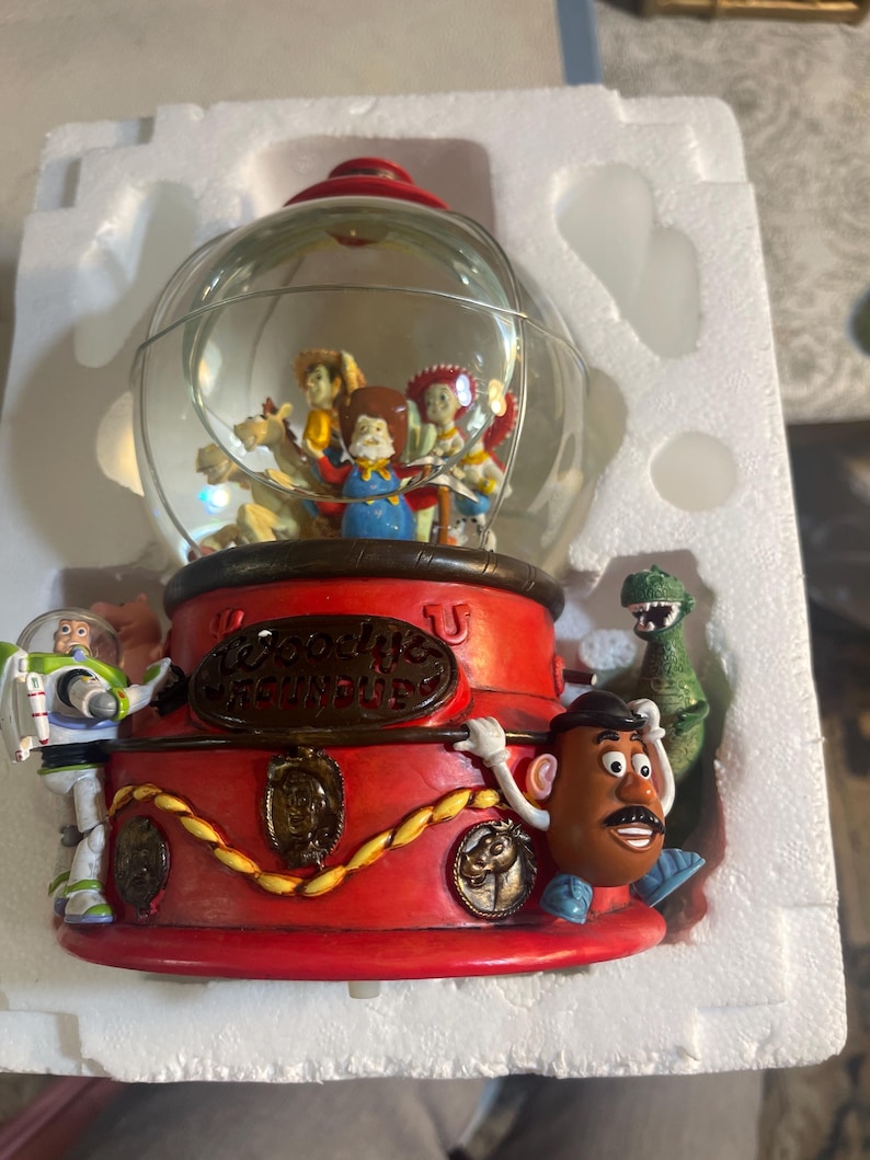May include: A Toy Story themed snow globe featuring Woody, Buzz Lightyear, Mr. Potato Head, Rex, and other characters. The base is red with gold accents and the text "Woody's Roundup". The globe is clear with a red top.