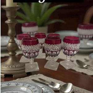 May include: An elegant table setting featuring six wine glasses with ruby red bowls and diamond-cut crystal bases. The word "Elegance" is displayed above the glasses. The table also includes plates, silverware, and a candle holder.