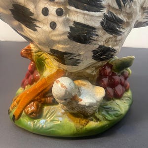 Pode incluir: Estatueta de cer&acirc;mica de uma galinha com um pintinho, pousada sobre uma base com uvas e folhas. A galinha tem um padr&atilde;o salpicado em branco e preto, e o pintinho &eacute; branco. A base &eacute; verde, com uvas vermelhas e galhos marrons.
