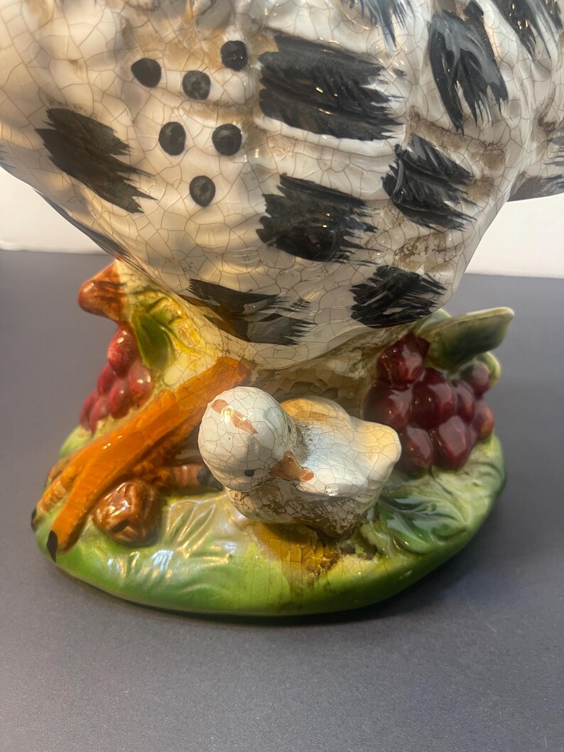 Pode incluir: Estatueta de cer&acirc;mica de uma galinha com um pintinho, pousada sobre uma base com uvas e folhas. A galinha tem um padr&atilde;o salpicado em branco e preto, e o pintinho &eacute; branco. A base &eacute; verde, com uvas vermelhas e galhos marrons.