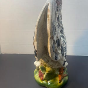 Pode incluir: Estatueta de galo de cer&acirc;mica com um design detalhado. O galo tem um corpo branco e cinza com manchas pretas, uma crista vermelha e uma base verde com detalhes vermelhos e brancos. As asas est&atilde;o abertas.
