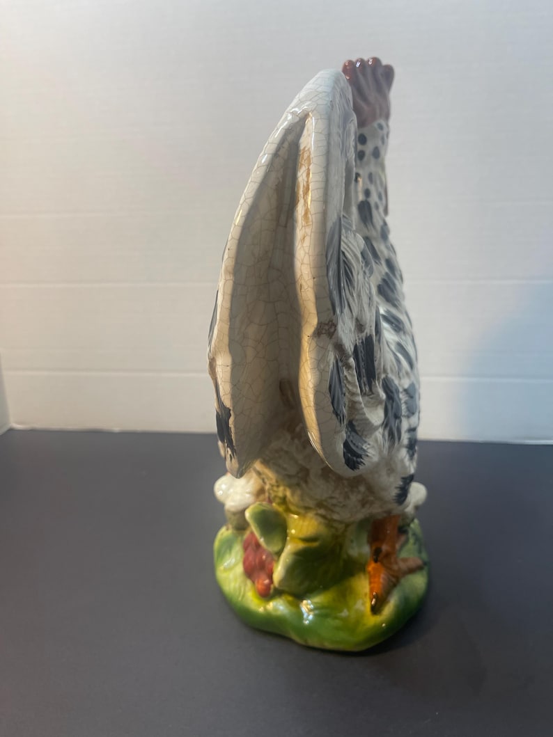 Pode incluir: Estatueta de galo de cer&acirc;mica com um design detalhado. O galo tem um corpo branco e cinza com manchas pretas, uma crista vermelha e uma base verde com detalhes vermelhos e brancos. As asas est&atilde;o abertas.