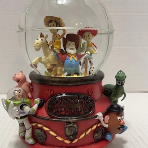 May include: A red and clear Toy Story-themed snow globe featuring characters like Woody, Buzz Lightyear, and Mr. Potato Head. The base is red with the text "Woody's Roundup". The globe is filled with figurines.