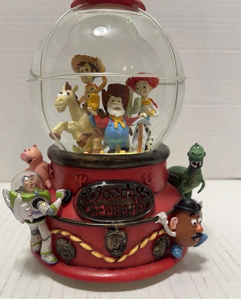 May include: A red and clear Toy Story-themed snow globe featuring characters like Woody, Buzz Lightyear, and Mr. Potato Head. The base is red with the text "Woody's Roundup". The globe is filled with figurines.