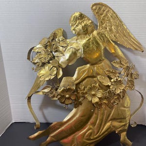 May include: A golden metal angel sculpture with outstretched wings and flowing robes. The angel is adorned with floral accents and holds a bouquet of flowers. The sculpture has a vintage, antique appearance.