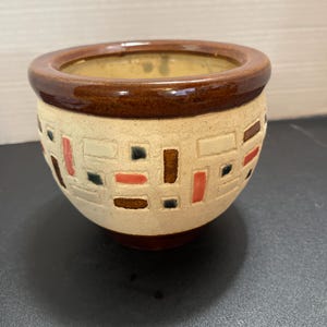 May include: A ceramic planter with a brown rim and base, and a beige body. The body features a geometric pattern of rectangles in red, brown, and black. The interior is a light yellow color.