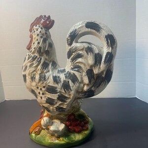 Pode incluir: Estatueta de galo de cer&acirc;mica com corpo creme, marcas pretas e crista vermelha. O galo est&aacute; sobre uma base verde com um pintinho e bagas vermelhas. A estatueta &eacute; um item decorativo.