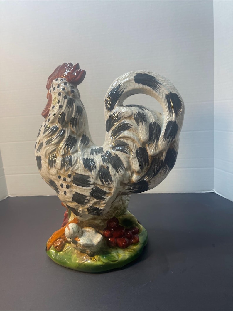 Pode incluir: Estatueta de galo de cer&acirc;mica com corpo creme, marcas pretas e crista vermelha. O galo est&aacute; sobre uma base verde com um pintinho e bagas vermelhas. A estatueta &eacute; um item decorativo.